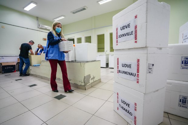 Sala com várias caixas térmicas brancas empilhadas e caixas de papelão organizadas. Algumas pessoas, usando máscaras, manuseiam as caixas.