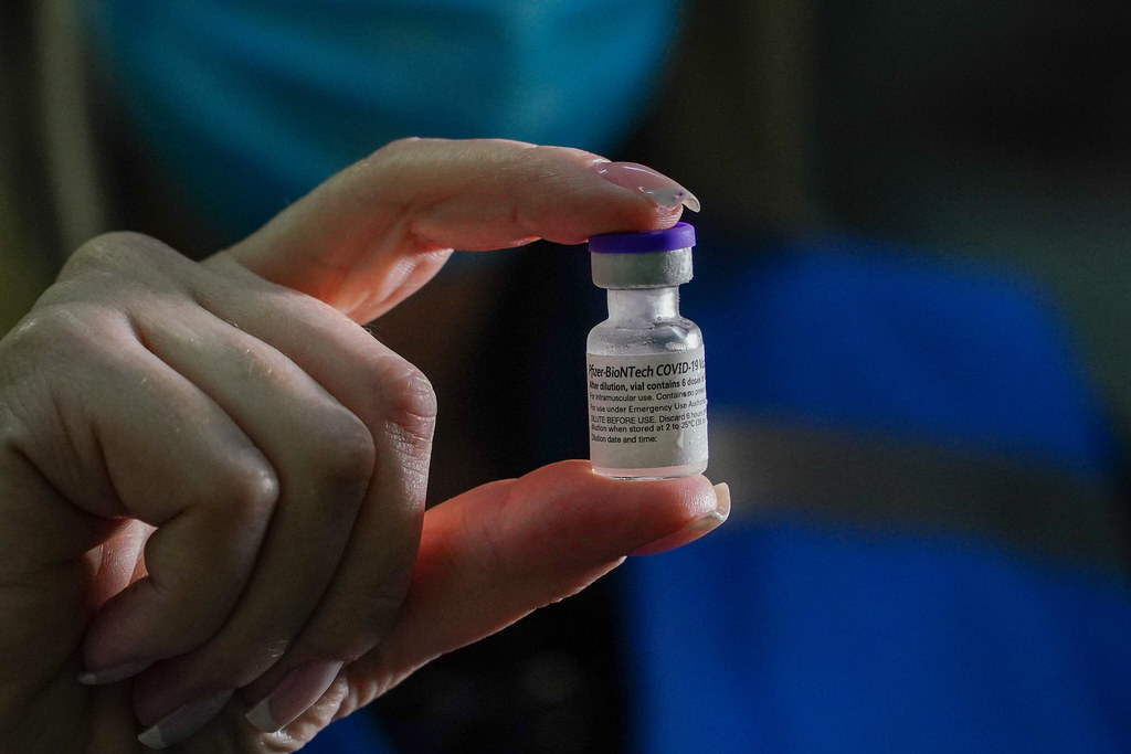 Mão feminina com unhas compridas e bem cuidadas segurando um frasco da vacina da Pfizer-BioNTech contra a COVID-19. O vidro transparente exibe o líquido do imunizante e o rótulo com a marca 'BioNTech' em destaque.