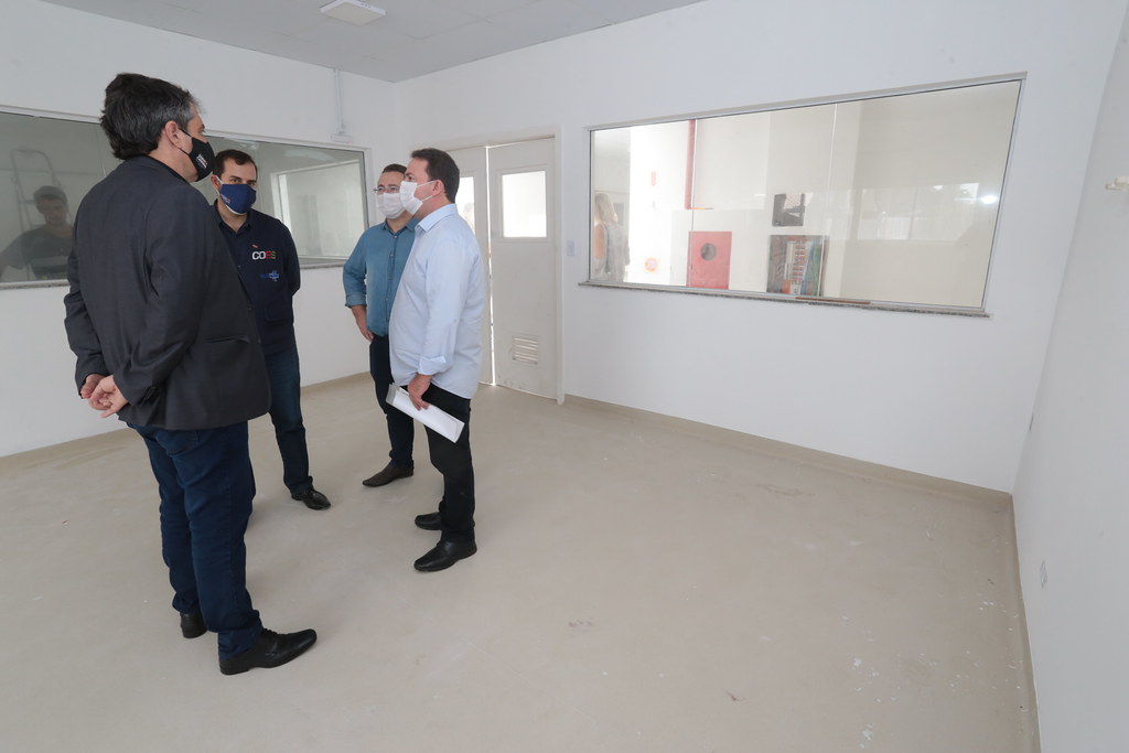 Quatro homens com máscaras conversam em uma sala branca e vazia com janelas de vidro; um deles segura documentos na mão.