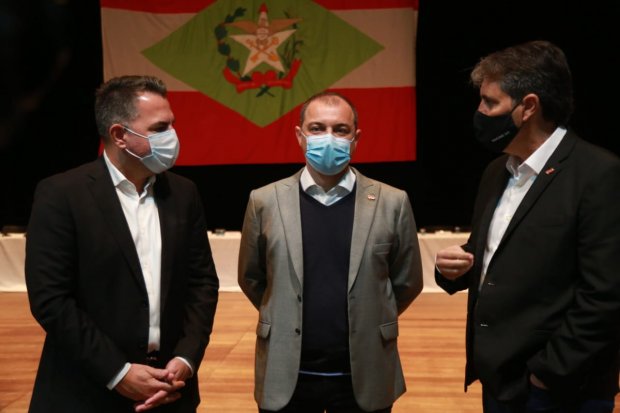 O governador de Santa Catarina conversa com duas pessoas em um auditório com paredes pretas e piso de madeira, durante reunião sobre a priorização da vacinação de profissionais da educação em 2021.