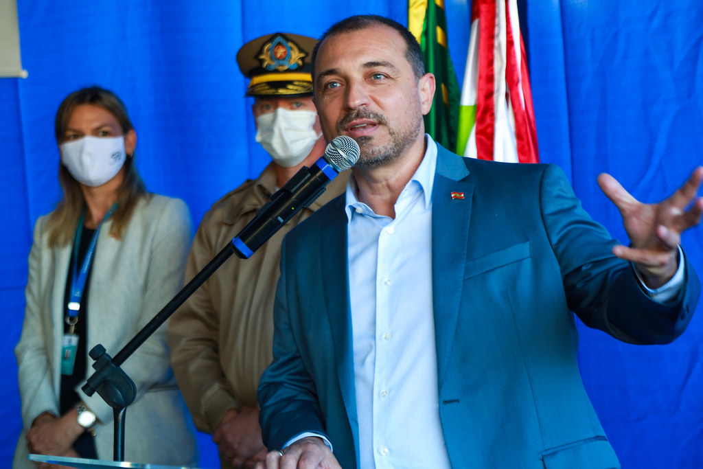 O governador de Santa Catarina, vestindo terno azul e broche com a bandeira do estado, fala em um microfone durante cerimônia oficial. Ao fundo, as bandeiras do Brasil e de Santa Catarina estão posicionadas, acompanhadas por uma mulher e um homem.
