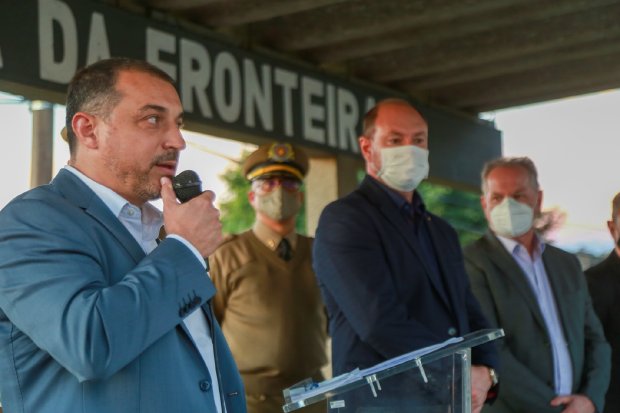 Governador Carlos Moisés discursando com microfone, acompanhado por autoridades mascaradas ao fundo, em evento que liberou R$ 6 milhões para o Hospital de São Miguel do Oeste e entregou viaturas à Polícia Militar. Placa parcialmente visível ao fundo com texto '...da fronteira'.