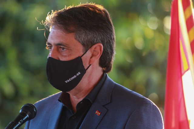 homem de terno azul e mascara preta discursando no microfone, ao fundo bandeira vermelha e amarela e árvores.