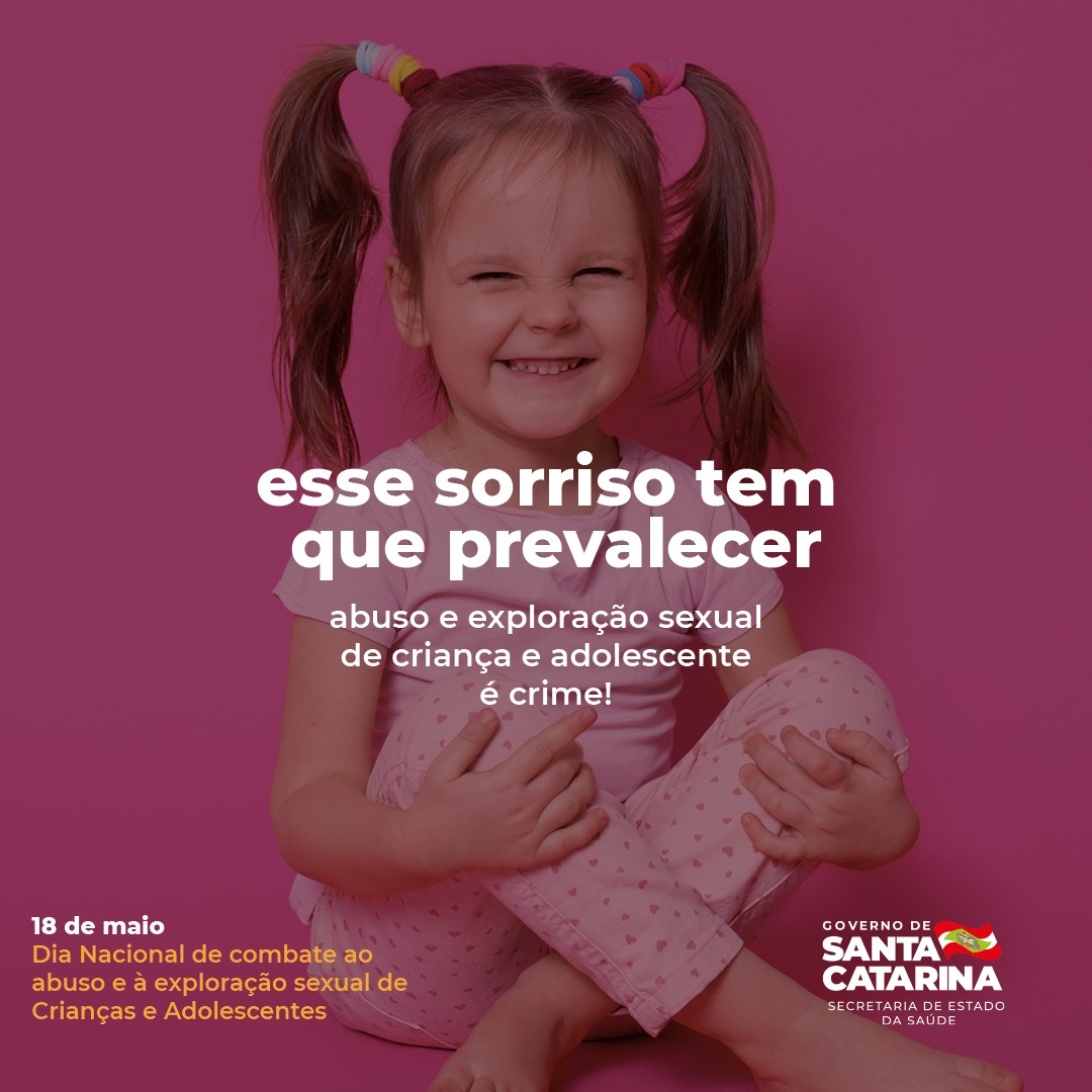 Professora lecionando em sala de aula para crianças pequenas. A sala possui chão de madeira, carteiras e ventilador.