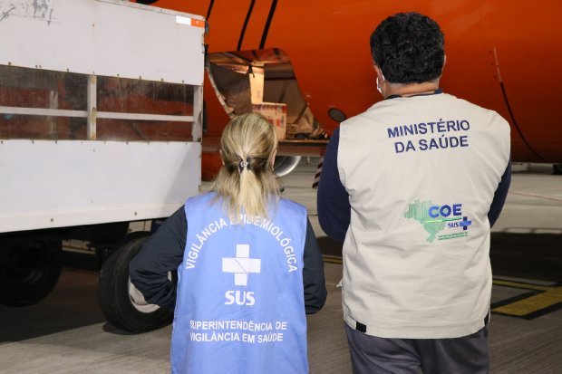 Mulher loira com colete azul da vigilância epidemiológica e a sua esquerda homem moreno do ministério da saúde acompanhando a retirada de caixas térmicas do avião.