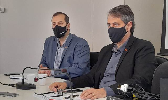 Dois homens com máscaras pretas de cabelo escuro sentados numa mesa com microfones em suas frentes