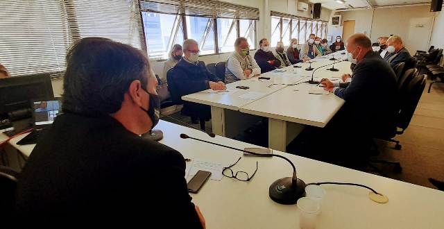 Reunião com cerca de 15 pessoas sentadas ao redor de duas mesas brancas unidas, em uma sala ampla com janelas laterais. Todos usam máscaras faciais e estão voltados para o centro da mesa, onde há microfones, copos plásticos e documentos. A maioria dos participantes aparenta estar em trajes formais, e o ambiente sugere uma discussão institucional ou governamental.