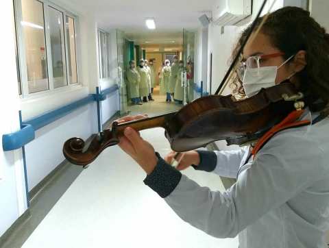 Mulher de óculos e de máscara tocando violino em corredor de hospital com médicos assistindo.