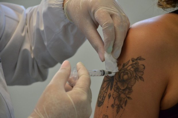 Mão de profissional da saúde com luva branca, aplicando vacina em um braço com tatuagem de flor no ombro