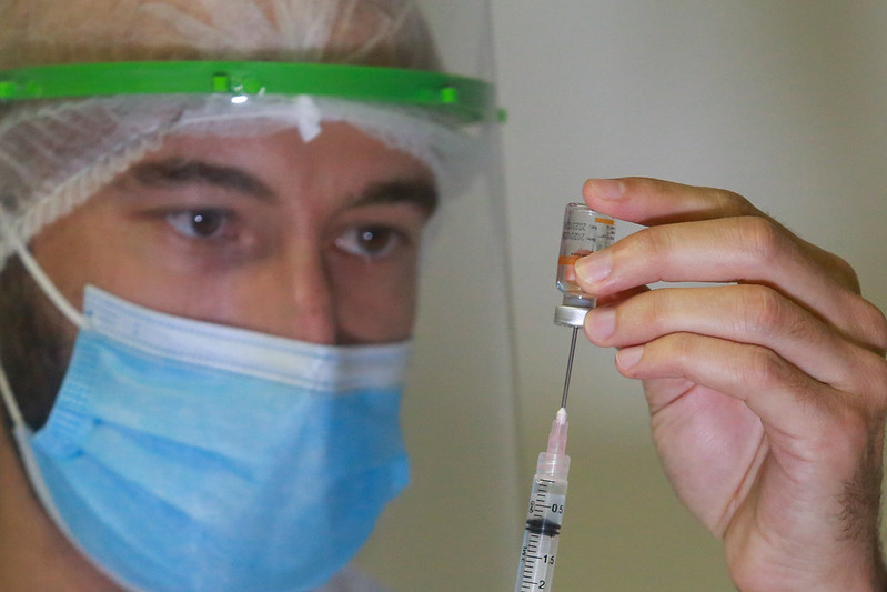 Homem profissional da saúde com máscara azul com um mão segurando um frasco e com a outra inserindo a seringa 