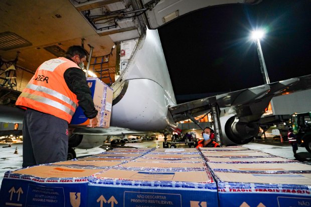 Caixas térmicas sendo descarregadas do avião. Dentro do avião existe uma pessoa mexendo em uma caixa, fora do avião duas pessoas estão carregando uma caixa enquanto uma terceira acompanha