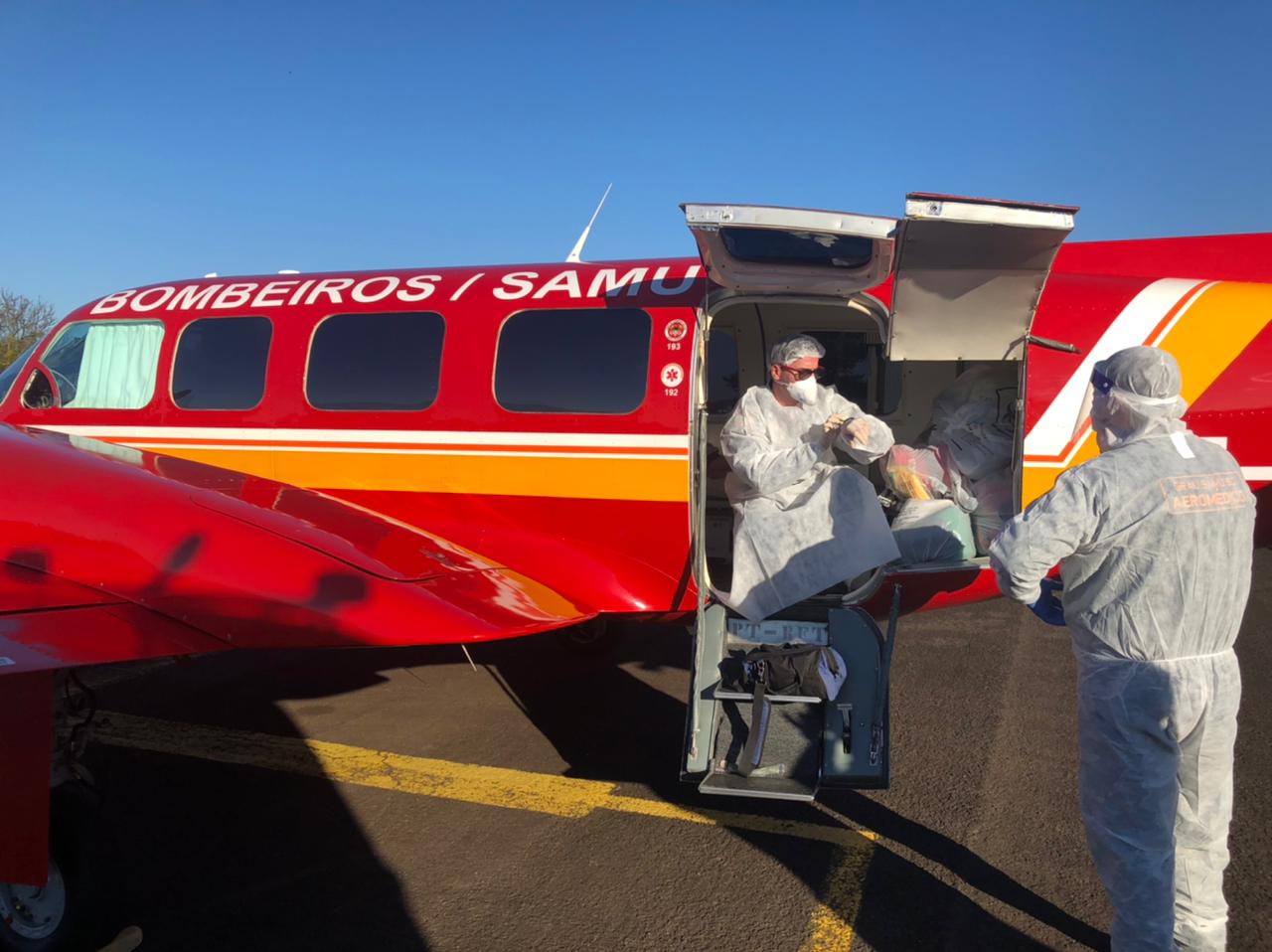 Pessoa com roupas de proteção saindo de avião todo vermelho dos bombeiros/samu.  Fora do avião tem outra pessoa com a mesma roupa esperando