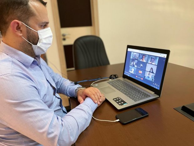 Homem de máscara sentado com braços apoiados numa mesa olhando para seu notebook que está com uma videochamada aberta.