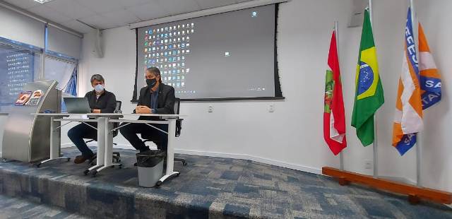 Dois homens sentados em cadeiras em cima do palco. Ao fundo existem 3 bandeiras, sendo uma a do Brasil, a outra de Santa Catarina. Ao fundo projetor reproduzindo área de trabalho de um computador.