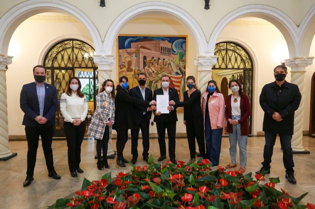 Grupo de onze pessoas usando máscaras posam para foto em ambiente interno com arquitetura clássica e arco central. Ao centro, três pessoas seguram um documento. Ao fundo, há uma pintura colorida de grande porte, e em primeiro plano, flores vermelhas decoram o local.