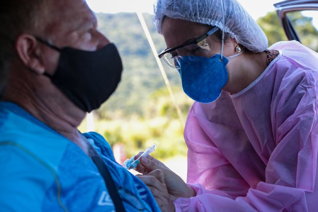 Profissional da saúde usando máscara azul, óculos protetor,  touca branca, e roupa rosa aplica dose de vacina em homem de roupa azul e máscara preta.