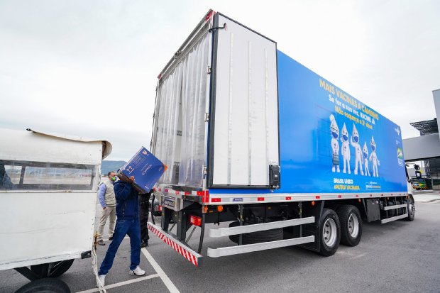 Homem todo de azul e tênis branco descarrega da caçamba de caminhão caixa azul contendo vacinas de COVID