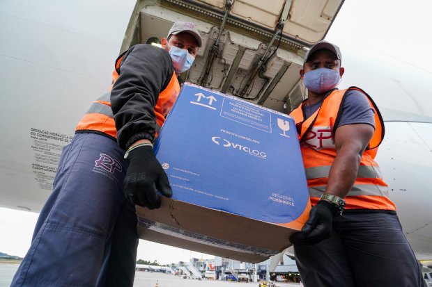 Dois homens com coletes laranja boné e máscara azul descarregam caixa