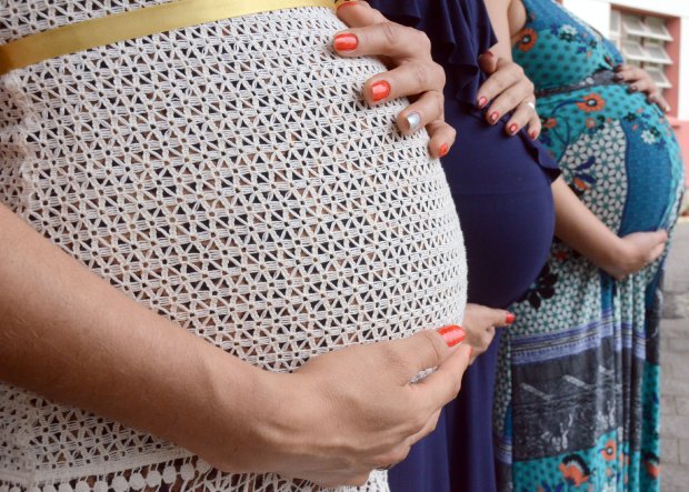 Três grávidas enfileiradas, o foco é na barriga delas, cada uma tem um vestido, o primeiro branco com bordados, o segundo azul e o terceiro verde e azul com estampas de flores.