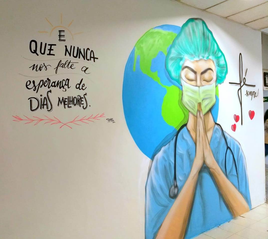 Mural em parede de hospital mostra profissional de saúde com jaleco azul, touca e máscara verde, de olhos fechados e mãos em posição de oração. Ao fundo, representação do planeta Terra. À esquerda, a frase: E que nunca nos falte a esperança de dias melhores. Ao lado da cabeça, a palavra fé com o "f" em forma de cruz e a palavra sempre, acompanhada de três corações vermelhos. Arte assinada por @sonhosdetinta.