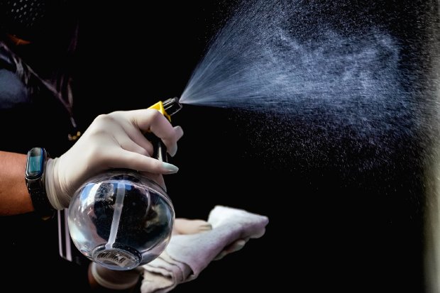 Mão segurando borrifador para desinfecção, espirrando líquido sanitizante, representando os protocolos de higiene adotados durante o primeiro ano de pandemia em Santa Catarina.