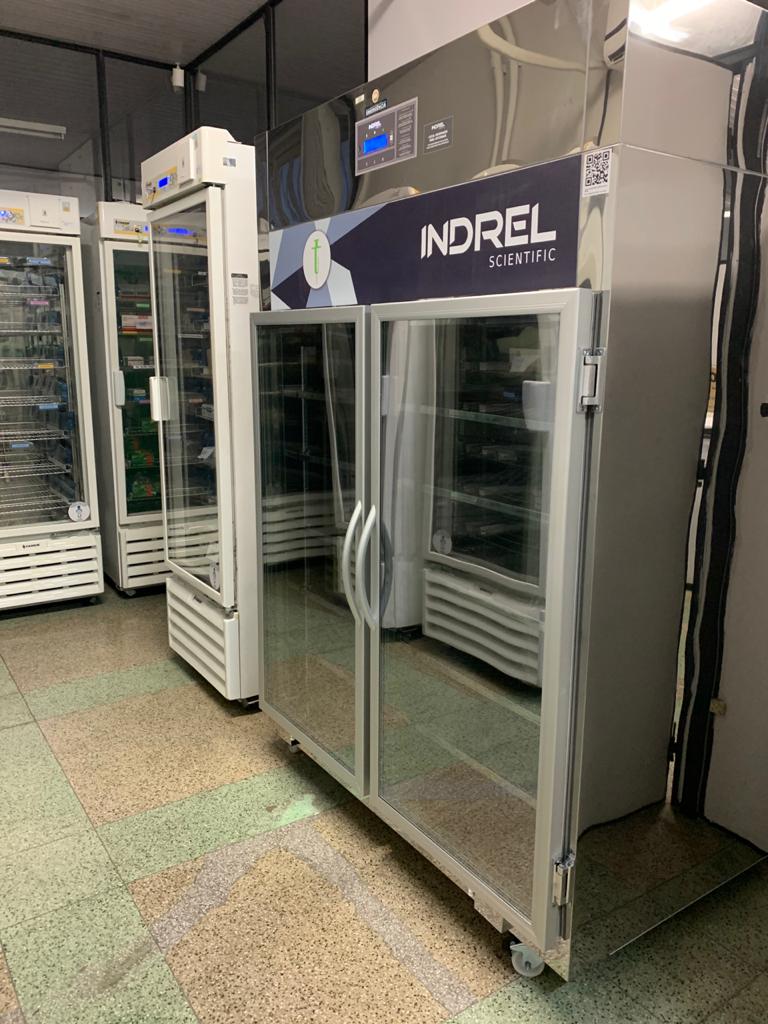 Câmaras frias para armazenamento de vacinas, com modelo cromado da Indrel Scientific em primeiro plano e unidades brancas ao fundo, recebidas pelo governo de Santa Catarina para ampliar capacidade de conservação de imunizantes.