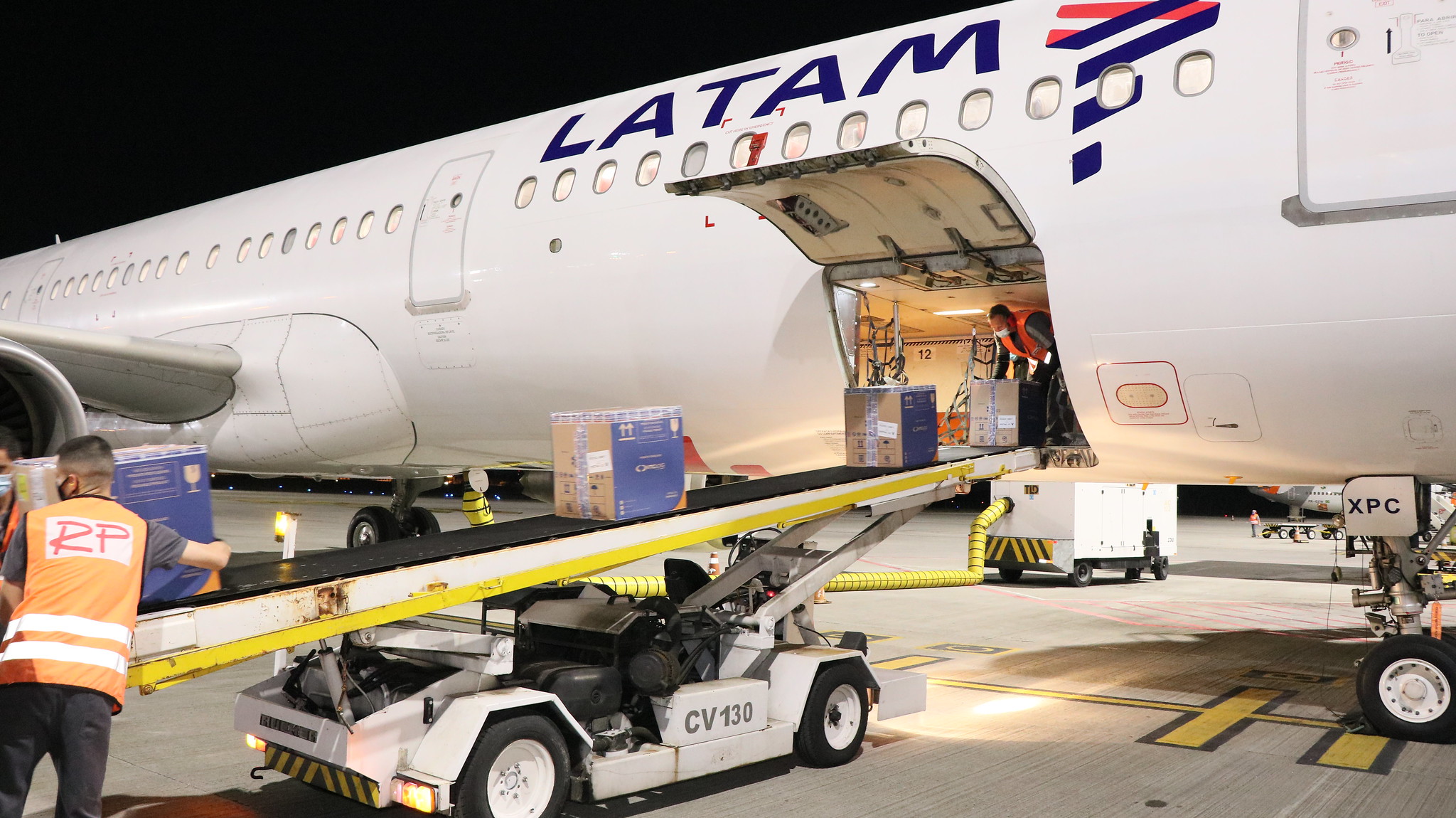 Caixas de vacina sendo descarregadas de avião latam branco por dois homens com coletes laranja