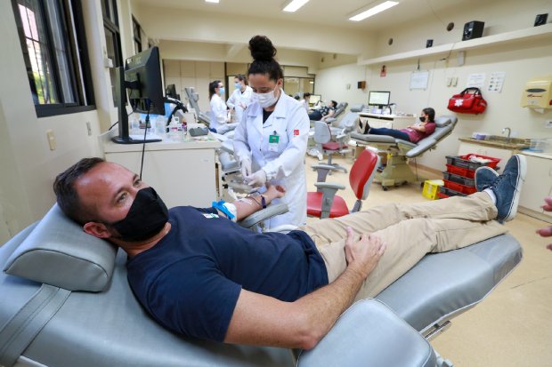 Homem sentado em poltrona doando sangue. Uma mulher de jaleco brando auxilia no procedimento. 