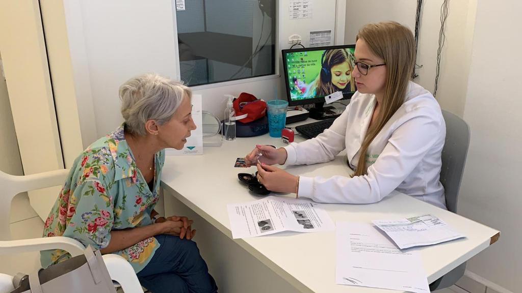 Profissional de saúde orienta uma mulher idosa durante atendimento em consultório. A paciente, de cabelos grisalhos, ouve atentamente enquanto a profissional, de jaleco branco, manuseia um aparelho auditivo. Sobre a mesa há papéis com informações e um computador com imagem de uma criança usando fones de ouvido.