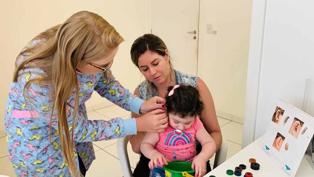 Profissional de saúde realiza exame na orelha de uma bebê sentada no colo da mãe, em ambiente clínico. A bebê segura um balde com peças coloridas enquanto a profissional, vestida com jaleco estampado com dinossauros, manuseia o equipamento auditivo. Sobre a mesa ao lado, há ilustrações mostrando diferentes formatos de orelha.