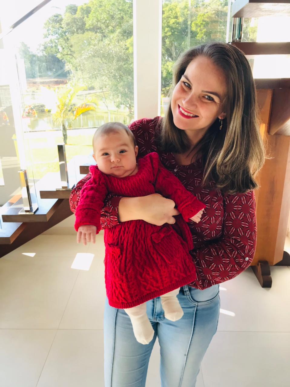 Mulher com roupa vermelha e calça jeans segurando bebê com vestido vermelho em interior de casa