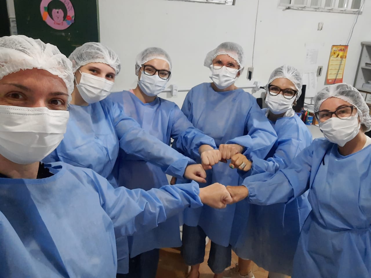 Seis profissionais de saúde mulheres uniformizadas (jaleco azul, touca e máscara) unindo os punhos em gesto de solidariedade, simbolizando a equipe no combate à COVID-19 em Santa Catarina.