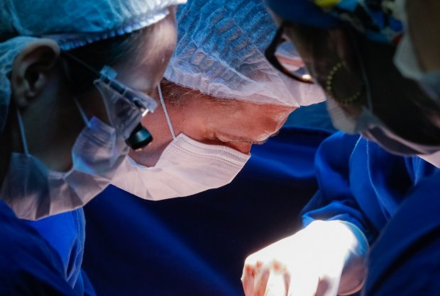 Três rostos de médicos prestando atenção em uma cirurgia, todos estão de máscara e touca e estão vestindo roupas azuis 