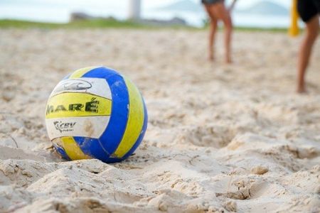 Bola de vôlei azul, amarela e branca com a palavra 'MARÉ' em destaque, apoiada na areia de uma praia. Ao fundo, pernas de pessoas em trajes esportivos sugerem um jogo de vôlei de praia em andamento.