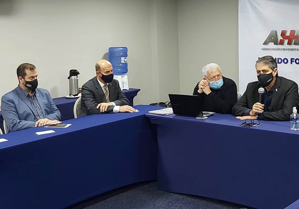 4 homens sentadas em mesa com toalha azul, um deles segura um microfone