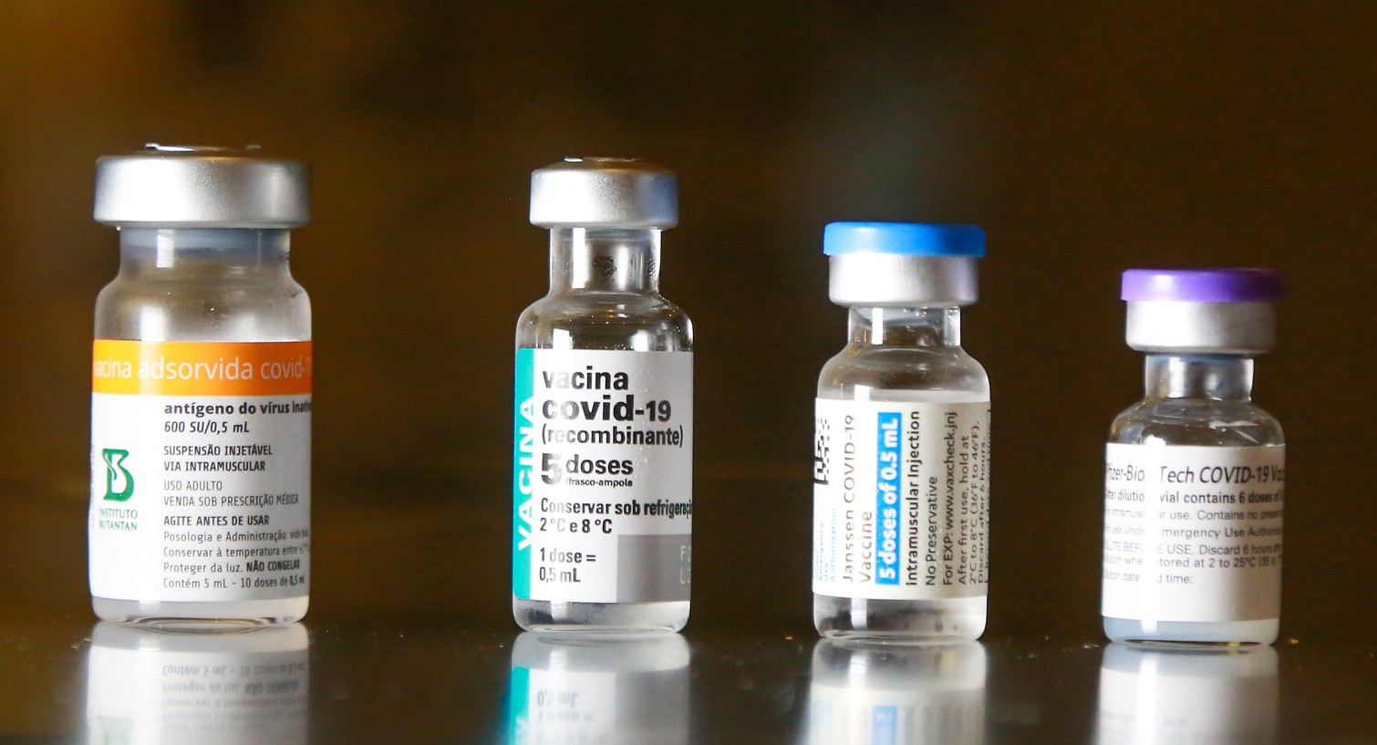 Quatro frascos da vacina Covishield (ChAdOx1 nCoV-19), desenvolvida pela AstraZeneca em parceria com a Universidade de Oxford, estão posicionados lado a lado sobre uma superfície branca. Cada frasco tem tampa verde e rótulo verde com informações sobre o imunizante.