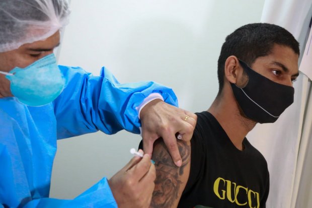Jovem com ombro tatuado com linhas pretas, camisa e máscara preta recebe vacina de profissional de saúde com roupa e máscara azul