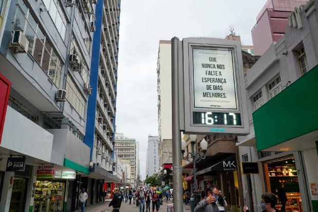 Rua movimentada no centro da cidade com pedestres circulando e relógio público marcando entre 16h e 17h, retratando o cenário urbano após alterações nas medidas sanitárias contra COVID-19 em Santa Catarina.
