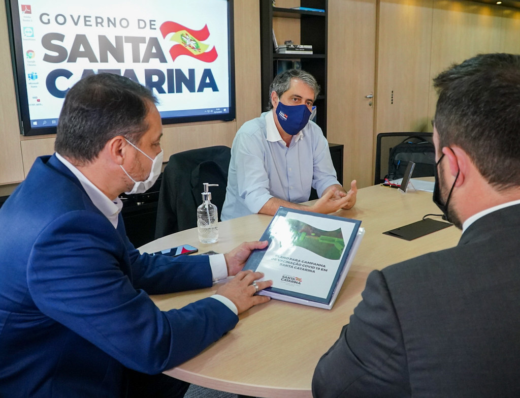 Reunião entre autoridades do Governo de Santa Catarina sobre a campanha de vacinação contra a COVID-19. Três homens com máscara estão sentados ao redor de uma mesa, analisando um documento com o título Plano para Campanha de Vacinação COVID-19 em Santa Catarina. Ao fundo, uma tela exibe o logotipo do Governo de Santa Catarina. Um frasco de álcool em gel está sobre a mesa, destacando os cuidados sanitários.