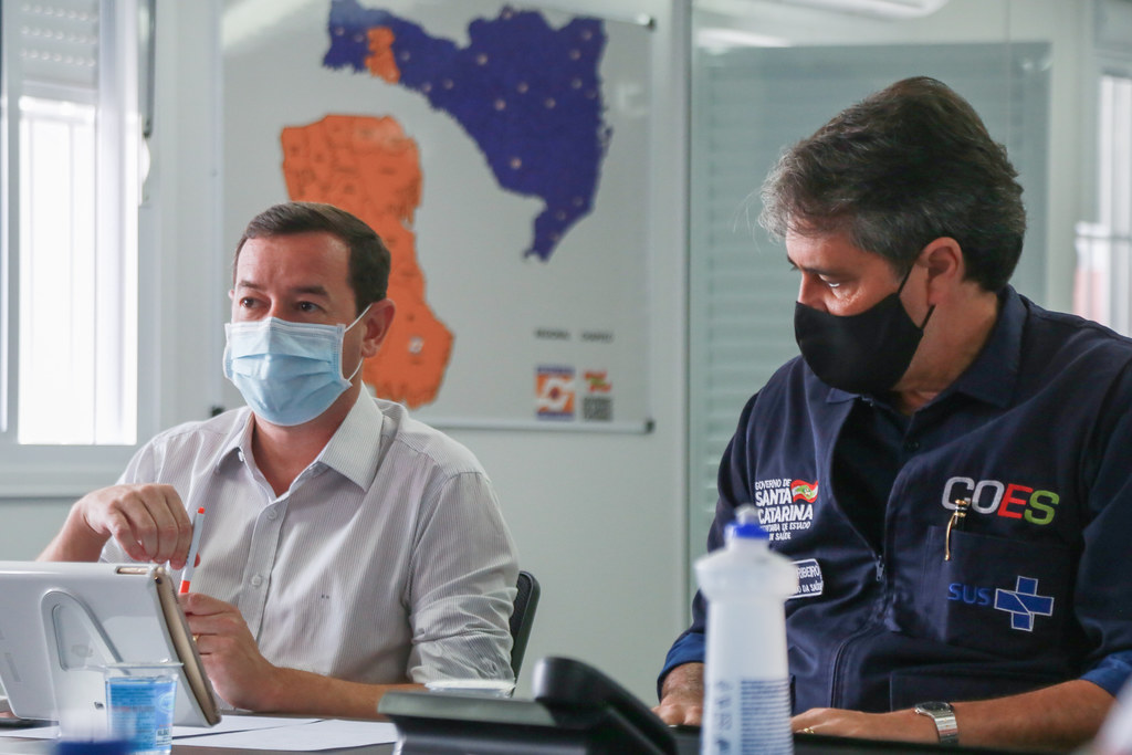 Dois homens sentados à mesa durante reunião; ambos usam máscaras. Um deles está com uma caneta na mão e tablet à frente. O outro veste colete com os logos do Governo de Santa Catarina, COES e SUS. Ao fundo, há um mapa do estado de Santa Catarina com áreas destacadas em laranja e azul.