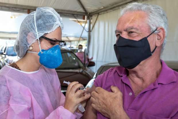 Profissional de saúde com roupa rosa, máscara azul e touca branca aplicando dose em idoso com roupa rosa e máscara preta