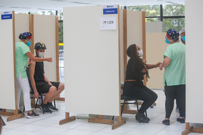 Profissionais de saúde aplicam vacinas contra a COVID-19 em pacientes dentro de cabines separadas por divisórias. Um dos pacientes usa capacete de ciclista e outro está com o braço estendido para receber a injeção. As áreas estão identificadas com placas, incluindo uma com o nome 'PFIZER'. Todos usam máscaras de proteção e o ambiente é bem iluminado, com janelas ao fundo.