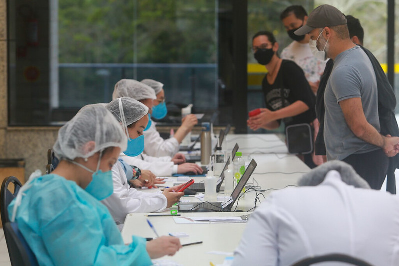 Profissionais da saúde com máscaras e toucas descartáveis atendem pessoas em uma mesa longa equipada com notebooks e materiais de registro. Os pacientes, também de máscara, aguardam em fila enquanto fornecem informações. A cena ocorre em um ambiente interno, com janelas de vidro ao fundo e iluminação natural.