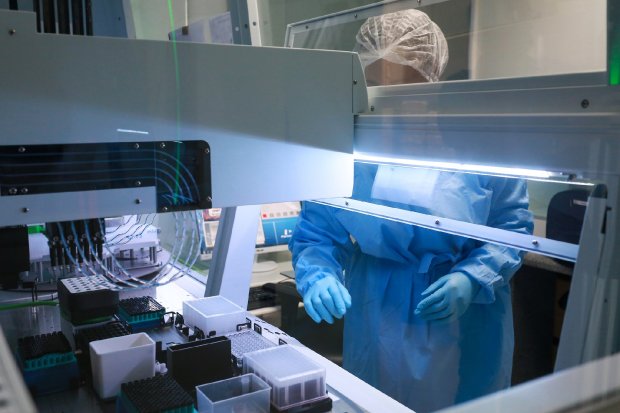 Profissional da saúde utilizando jaleco, touca e luvas em ambiente laboratorial, operando equipamento automatizado de análise com múltiplos compartimentos e tubos, sob luz artificial intensa.