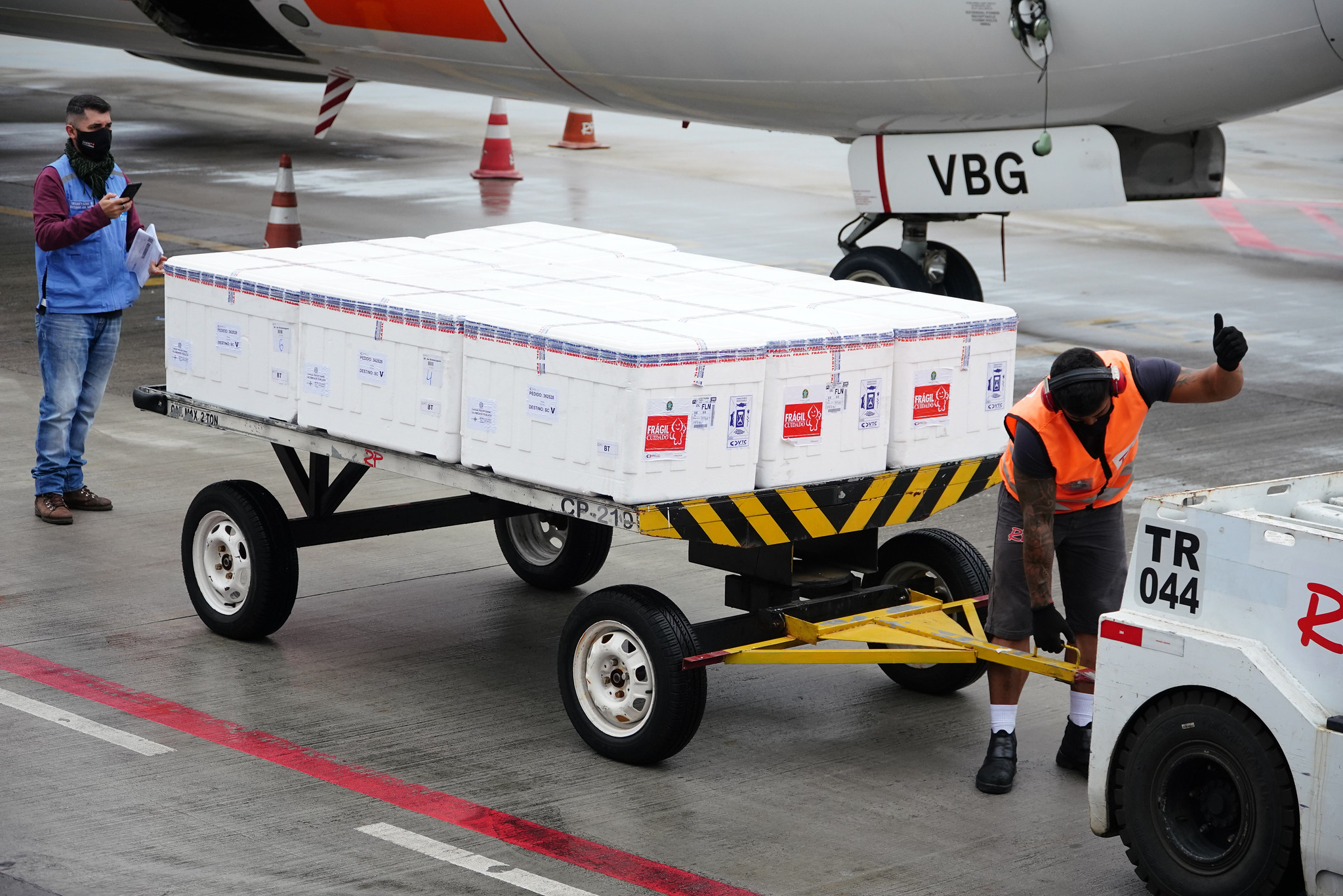 Caixas empilhadas em estrutura com quatro rodas sendo conectada por homem de colete laranja a um carro no aeroporto. Ao fundo Conseguimos ver um avião.