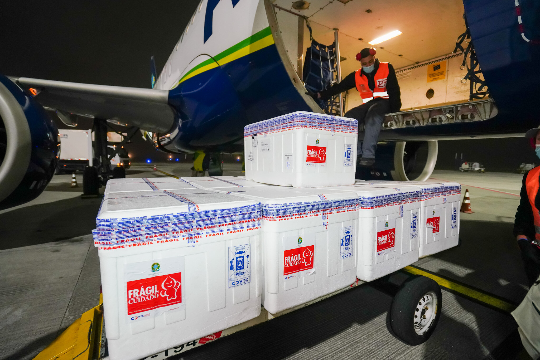 Caixas brancas em carrinho no aeroporto de Florianópolis anoite. Ao fundo tem um avião com homem de colete laranja dentro.