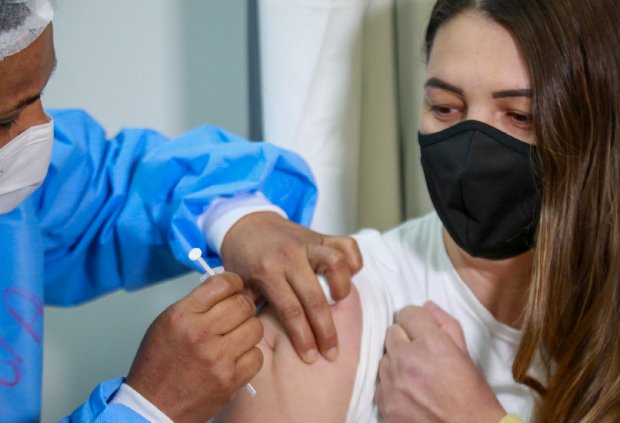 Mulher com roupa branca e máscara preta recendo vacina no ombro aplicada por mulher de roupa azul e máscara branca