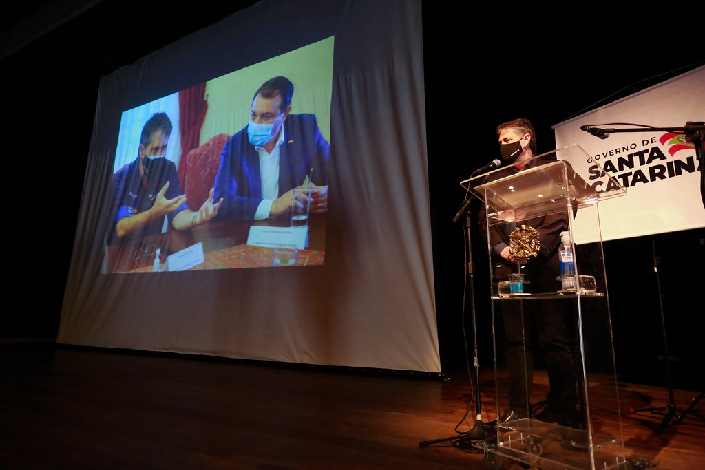 Pessoa discursando no palco, ao seu fundo tem um projetor exibindo uma foto de dois homens sentados