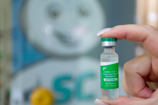 Mão com unhas compridas segurando frasco da vacina Covishield, com o personagem Zé Gotinha e a sigla 'SC' desfocados ao fundo.