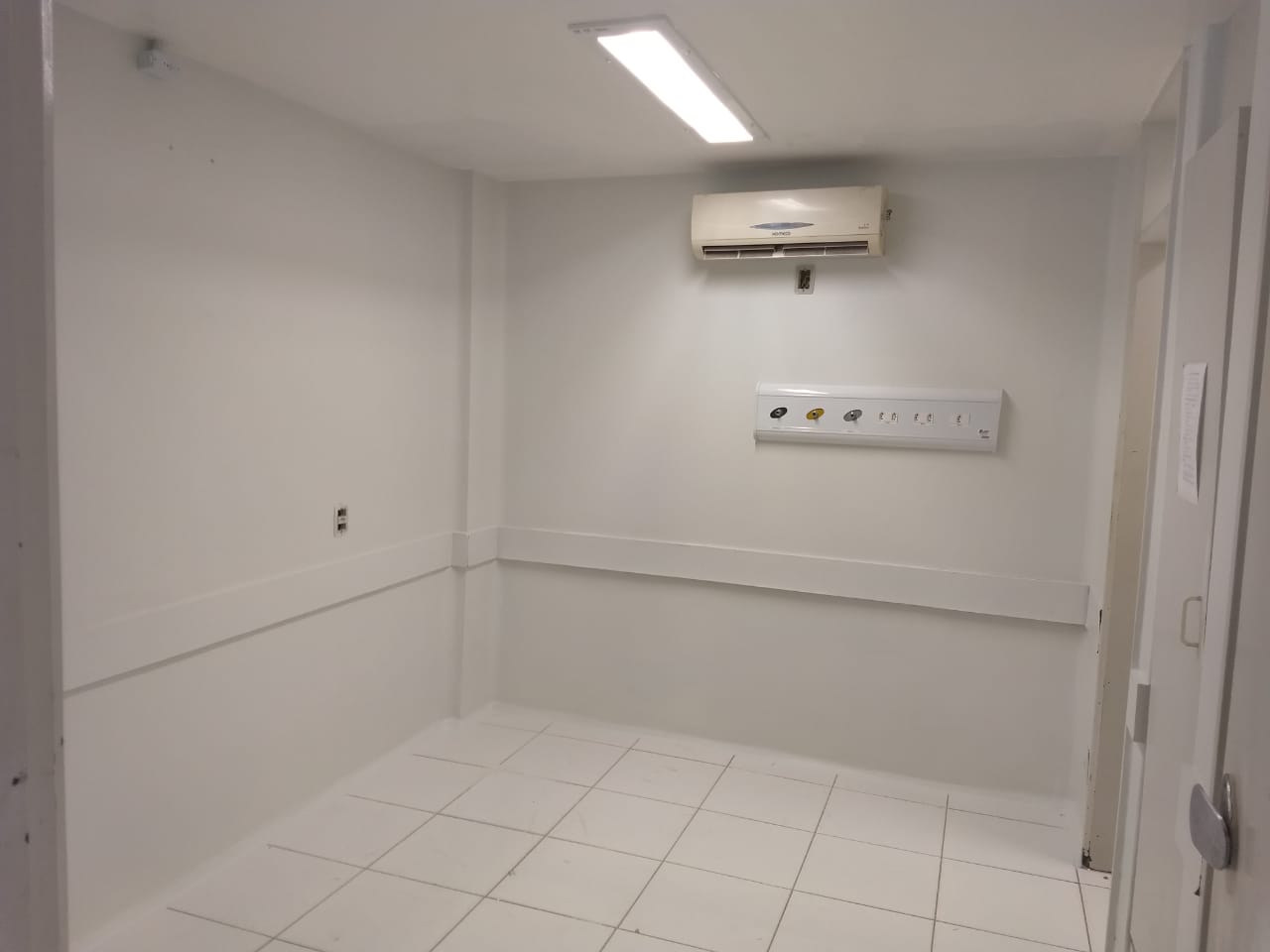 Área interna de hospital, paredes brancas e chão branco. No fundo tem um ar-condicionado.